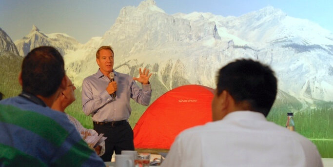 A man in a button-down shirt gives a speech to a room of executives.